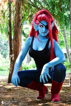 a woman with red hair and blue makeup sitting on the ground in front of trees