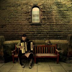 a person sitting on a bench with an accordion in front of a brick wall and window