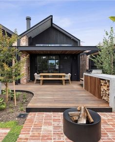 a wooden deck with benches and fire pit in front of a black house that is surrounded by brick pavers