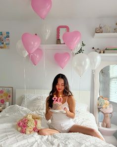 a woman is sitting on her bed with balloons and a cake in front of her