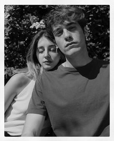 a man and woman sitting next to each other in front of some bushes with their eyes closed