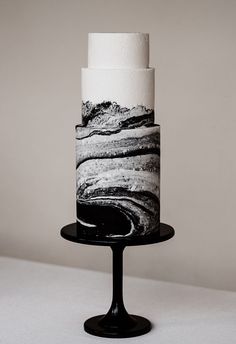 a three tiered cake with black and white icing on a stand in front of a wall