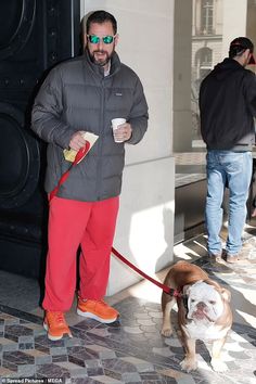 a man in grey jacket and red pants holding a coffee cup while standing next to a dog