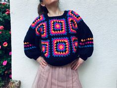 a woman standing in front of a white wall wearing a colorful crochet sweater