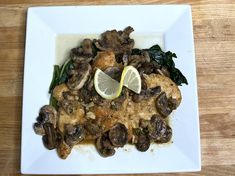 a white plate topped with meat covered in mushrooms and spinach next to a lemon wedge