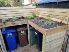 an outdoor garden area with various plants and trash cans in the corner, along side a wooden fence