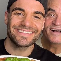 two men are smiling while holding a plate with food on it and one is looking at the camera