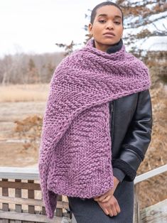 a woman wearing a purple knitted shawl standing on a wooden bench in front of a field