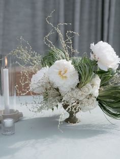 a vase filled with white flowers and greenery next to a candle on a table