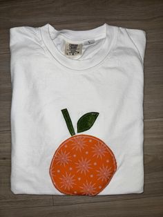 an orange t - shirt with a green leaf on the front and white back, sitting on a wooden floor