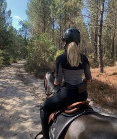 a woman riding on the back of a brown horse down a dirt road surrounded by trees