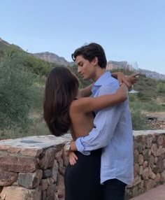 a man and woman standing next to each other on a stone wall with mountains in the background