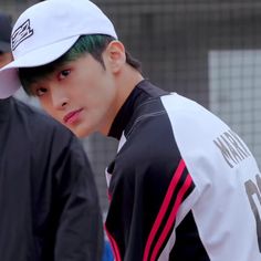 a young man with green hair wearing a white hat and black jacket, standing in front of a fence