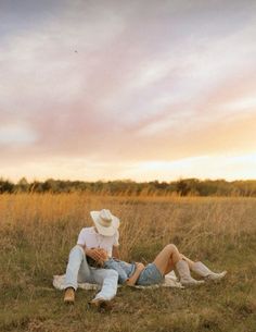 two people sitting on the ground in an open field at sunset, with one person wearing a cowboy hat