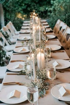 a long table with white plates and silverware on it, surrounded by greenery