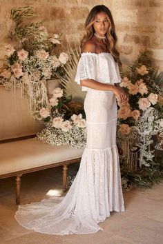 a woman standing in front of flowers wearing a white dress with off the shoulder sleeves