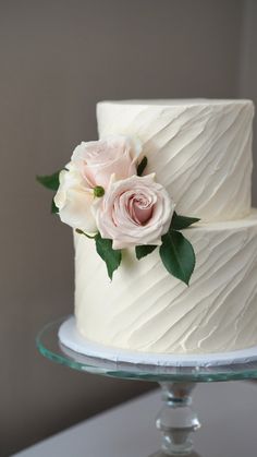 a three layer cake with white frosting and pink roses on the top is sitting on a glass plate