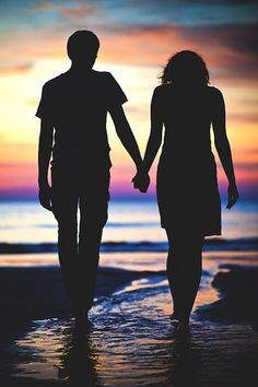 two people holding hands while walking on the beach at sunset or dawn with their arms around each other