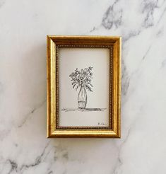 a gold frame holds a drawing of a vase with flowers in it on a marble surface