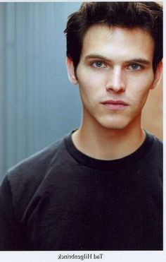 a young man with blue eyes and black t - shirt looking at the camera while standing in front of a wall