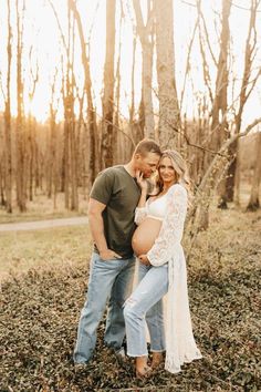 a pregnant couple standing next to each other in front of some trees and grass with the sun shining on them