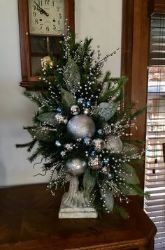 a christmas tree decorated with silver balls and greenery on a table in front of a clock