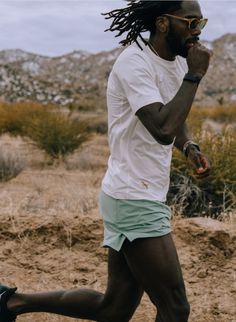 a man with dreadlocks running in the desert wearing green shorts and a white t - shirt