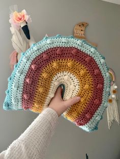 a person holding up a crocheted rainbow cushion in front of a gray wall