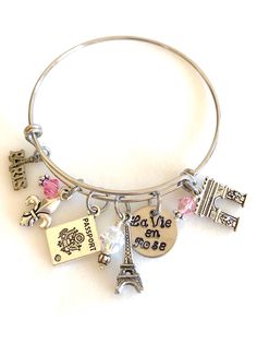 "Perfect souvenir of an enchanting trip to Paris, France. Adjustable silver bangle is highlighted with hand stamped quote \"La Vie en Rose\" and 5 detailed Parisian charms accented with rose colored Swarovski crystals AB. Adjustable bangle is high quality stainless steel and measures 2.5\" diameter to fit most. Hand stamped 5/8\" 18 gauge aluminum disc reads \"La Vie en Rose\". Two 6 mm rose colored Swarovski Crystals AB and one larger 8 mm clear Crystal AB accent the charms. Two-sided 3D pewter Silver Nickel-free Bracelet Souvenir, Nickel-free Silver Bracelets As Souvenir, Nickel-free Pink Bangle Charm Bracelet, Nickel Free Pink Bangle Charm Bracelet, Paris Charm Bracelet, Paris Bracelet, Paris Vacation, Paris Trip, Pink Swarovski