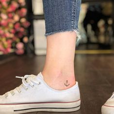 a small anchor tattoo on the foot of a woman's left ankle, while she is wearing jeans and white tennis shoes