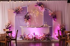 a purple and white wedding reception setup with flowers on the back wall, candles lit up in front of it