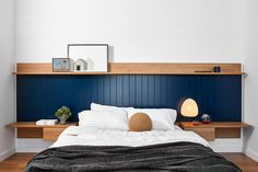 a bed with white sheets and blue headboard next to a wooden shelf on the wall