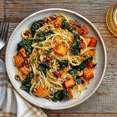 a white bowl filled with pasta and vegetables on top of a wooden table next to a glass of beer