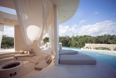 an outdoor lounge area next to a swimming pool with white drapes and pillows on it