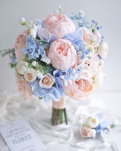 a bridal bouquet with blue and pink flowers on a white table cloth next to a card