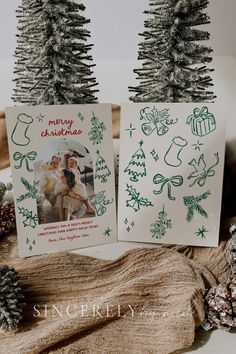 two christmas cards sitting on top of a table next to pine cones and fir trees