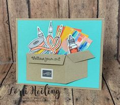 a card with an open cardboard box filled with art supplies on top of a wooden table