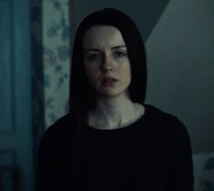 a woman with dark hair and blue eyes stares at the camera in a dimly lit room