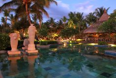 an outdoor swimming pool surrounded by palm trees and lights at dusk with tiki huts in the background