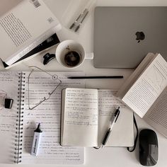 an open notebook, coffee cup and computer keyboard on a white desk with various items surrounding it