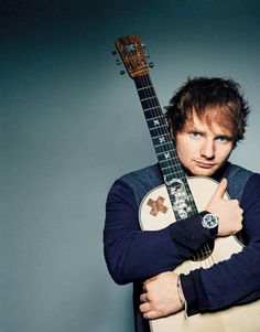 a man holding an acoustic guitar in his right hand and looking at the camera while wearing a blue sweater