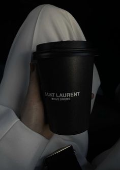 a person in white clothing holding a black cup with the word saint laurent on it