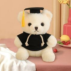 a white teddy bear wearing a graduation cap and gown sitting in front of a pink vase