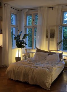 an unmade bed sitting in front of two windows next to a lamp and potted plant