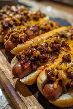 three hot dogs covered in chili and cheese sitting on a wooden cutting board next to other food items