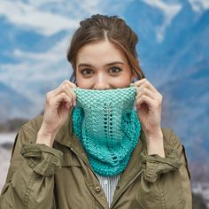 a woman holding up a knitted scarf to her face with mountains in the background