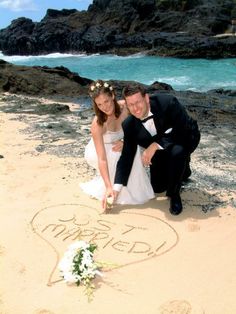 a newly married couple kneeling on the beach in front of their heart shaped wedding sign