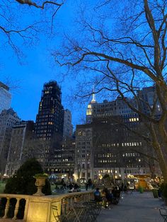 people sitting at tables in the middle of a park with tall buildings and trees on either side