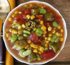 a white bowl filled with corn and vegetables on top of a wooden table next to a spoon