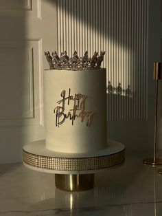 a white birthday cake sitting on top of a gold stand next to a wall and door